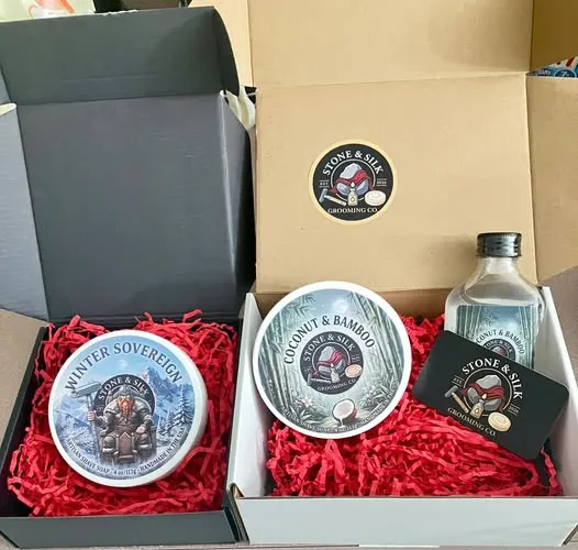 Product photography of Stone & Silk Grooming Co. shaving soap tubs, showing the brand's packaging and label artwork for its Coconut & Bamboo and Winter Sovereign scents.