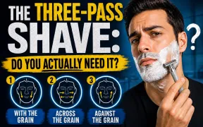 Man with dark hair and shaving cream on his cheek holding a chrome double-edge safety razor at his jawline, dark navy background, THE THREE-PASS SHAVE headline in white and yellow block letters with DO YOU ACTUALLY NEED IT below, three numbered face diagrams showing WITH THE GRAIN, ACROSS THE GRAIN, AGAINST THE GRAIN directions along the bottom