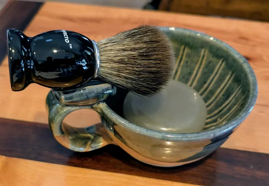A badger brush resting on the lip of a hand-thrown ceramic shave bowl glazed in green and cream with a thumb ring on the side, the bowl full of freshly whipped lather, sitting on a walnut and maple butcher block
