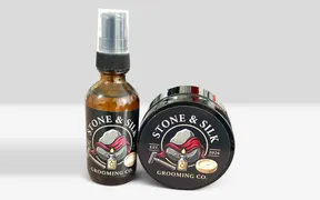 Stone and Silk Grooming Co pre-shave oil in an amber glass bottle with a pump top standing beside a black shave soap tub tipped on its side, both carrying the brand logo of a grey stone wrapped in red silk above a safety razor, an oil bottle and a soap puck