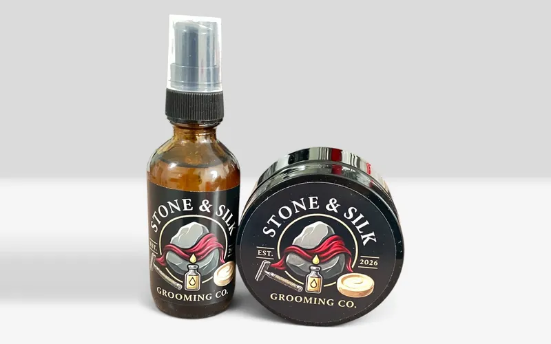 Stone and Silk Grooming Co pre-shave oil in an amber glass bottle with a pump top standing beside a black shave soap tub tipped on its side, both carrying the brand logo of a grey stone wrapped in red silk above a safety razor, an oil bottle and a soap puck