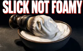 A shaving brush loaded with thick glossy white lather resting on the rim of a lather bowl on a dark slate counter, soft peaks of lather standing up, warm side-light, headline SLICK NOT FOAMY in white block letters with a red drop shadow across the top