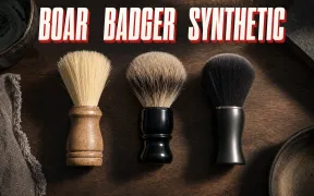 An overhead flatlay on a dark walnut counter of three shaving brushes standing in a row, a cream-bristled boar brush with a wooden handle, a soft gray and tan badger brush with a black resin handle, and a sleek modern synthetic brush with uniform dark fibers, headline BOAR BADGER SYNTHETIC in white block letters with a red drop shadow across the top