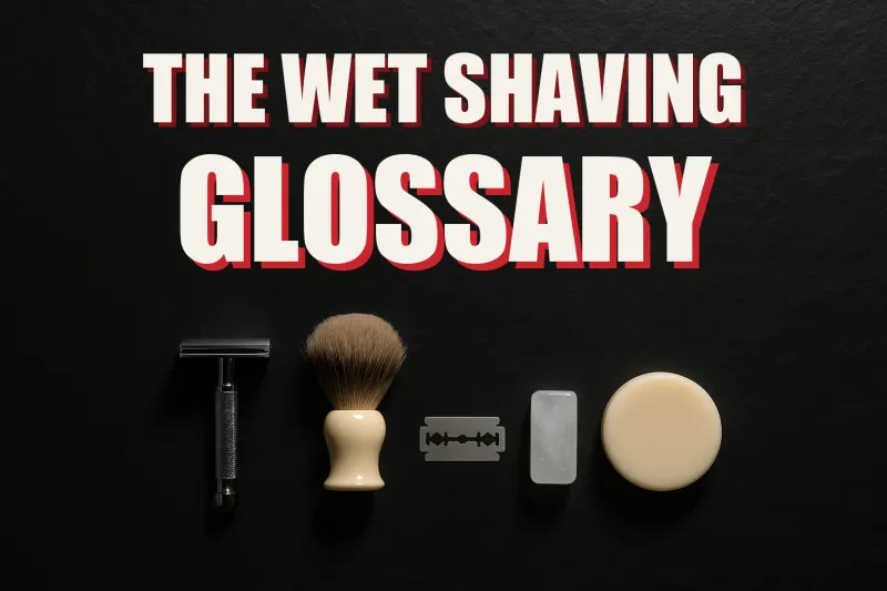 A flat lay of wet shaving gear arranged like a museum specimen board on dark slate, razor, brush, blade, soap puck and alum block each in its own pool of light, headline THE WET SHAVING GLOSSARY in white block letters with a red drop shadow