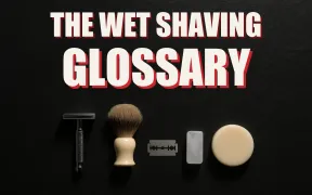 A flat lay of wet shaving gear arranged like a museum specimen board on dark slate, razor, brush, blade, soap puck and alum block each in its own pool of light, headline THE WET SHAVING GLOSSARY in white block letters with a red drop shadow
