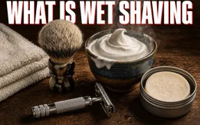 A warm overhead flatlay on dark walnut of a classic wet shaving kit, a chrome double-edge safety razor, a badger shaving brush standing beside a ceramic bowl crowned with thick white lather, a soap puck in a tin and a folded towel, headline WHAT IS WET SHAVING in white block letters with a red drop shadow across the top