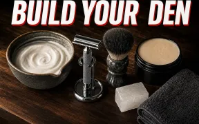 An overhead flatlay on dark walnut of a wet shaving station, a chrome safety razor resting on a stand, a shaving brush drying beside a ceramic lather bowl, a soap puck, an alum block and a folded towel under warm side light, headline BUILD YOUR DEN in white block letters with a red drop shadow across the top
