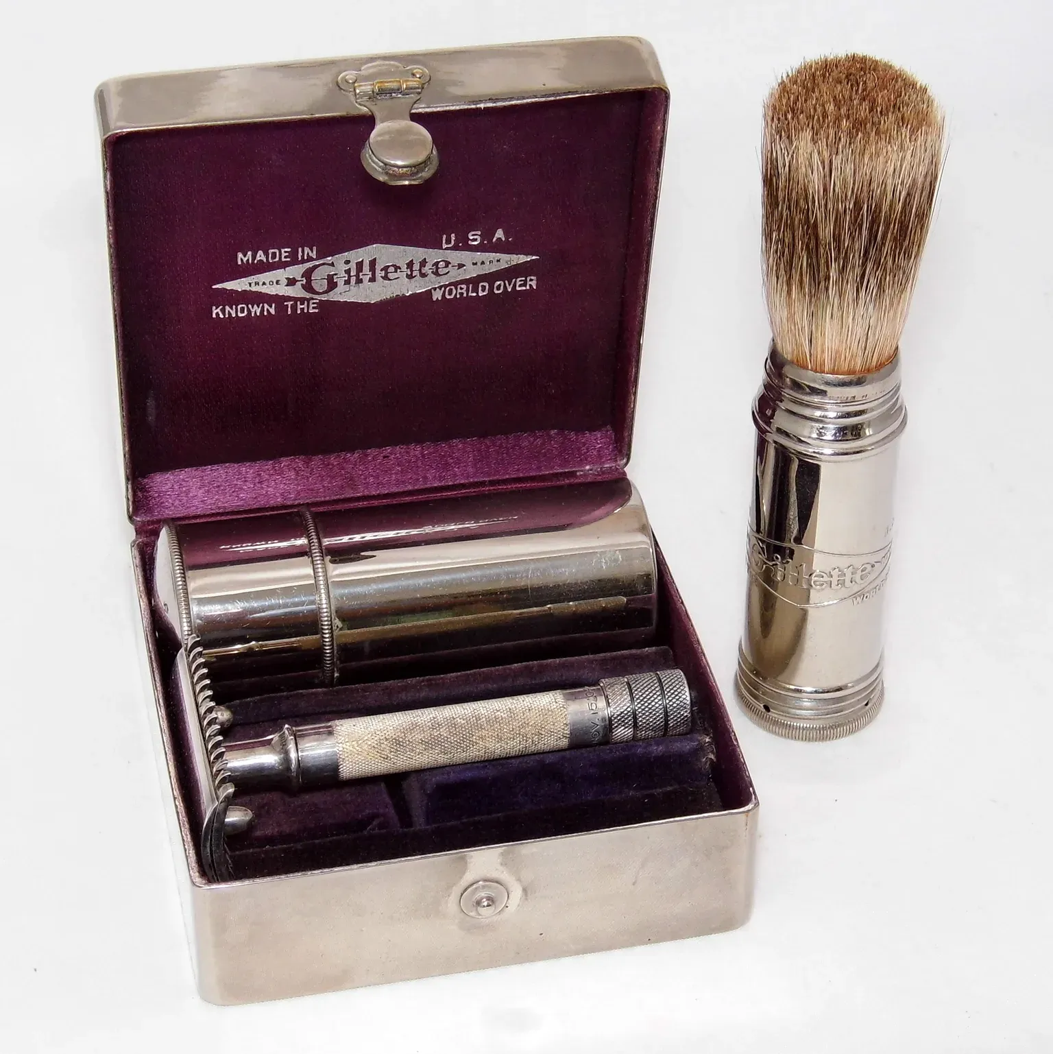 An open nickel plated Gillette Combination set with purple velvet lining, containing an Old Type three piece razor, a blade box and a travel shaving brush, with the Gillette diamond logo stamped inside the lid.