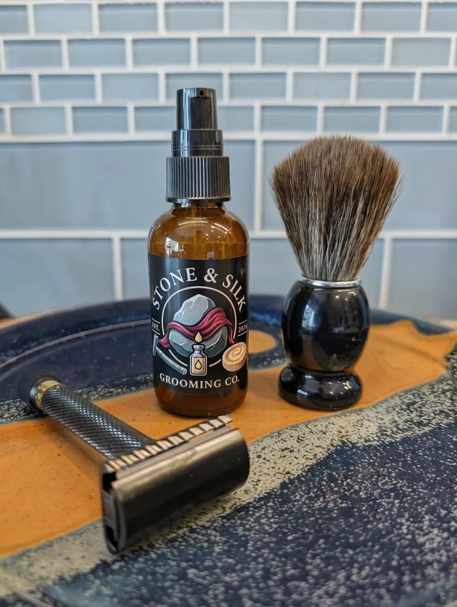 A Stone and Silk Grooming Co pre-shave oil bottle standing between a badger shave brush and a knurled double-edge safety razor, arranged on a blue and orange hand-glazed ceramic tray against a pale blue tiled wall