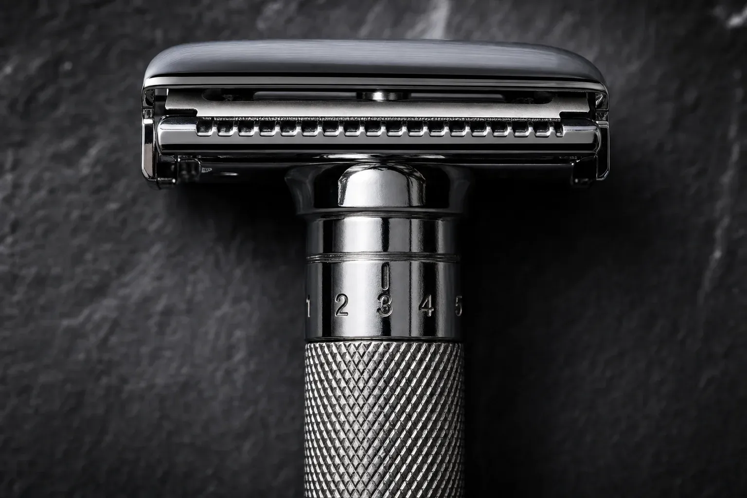 Macro photograph of an adjustable double-edge safety razor against dark slate, the knurled adjustment collar on the handle engraved with numbers one through five and an index line, beneath a chrome head with a scalloped safety bar