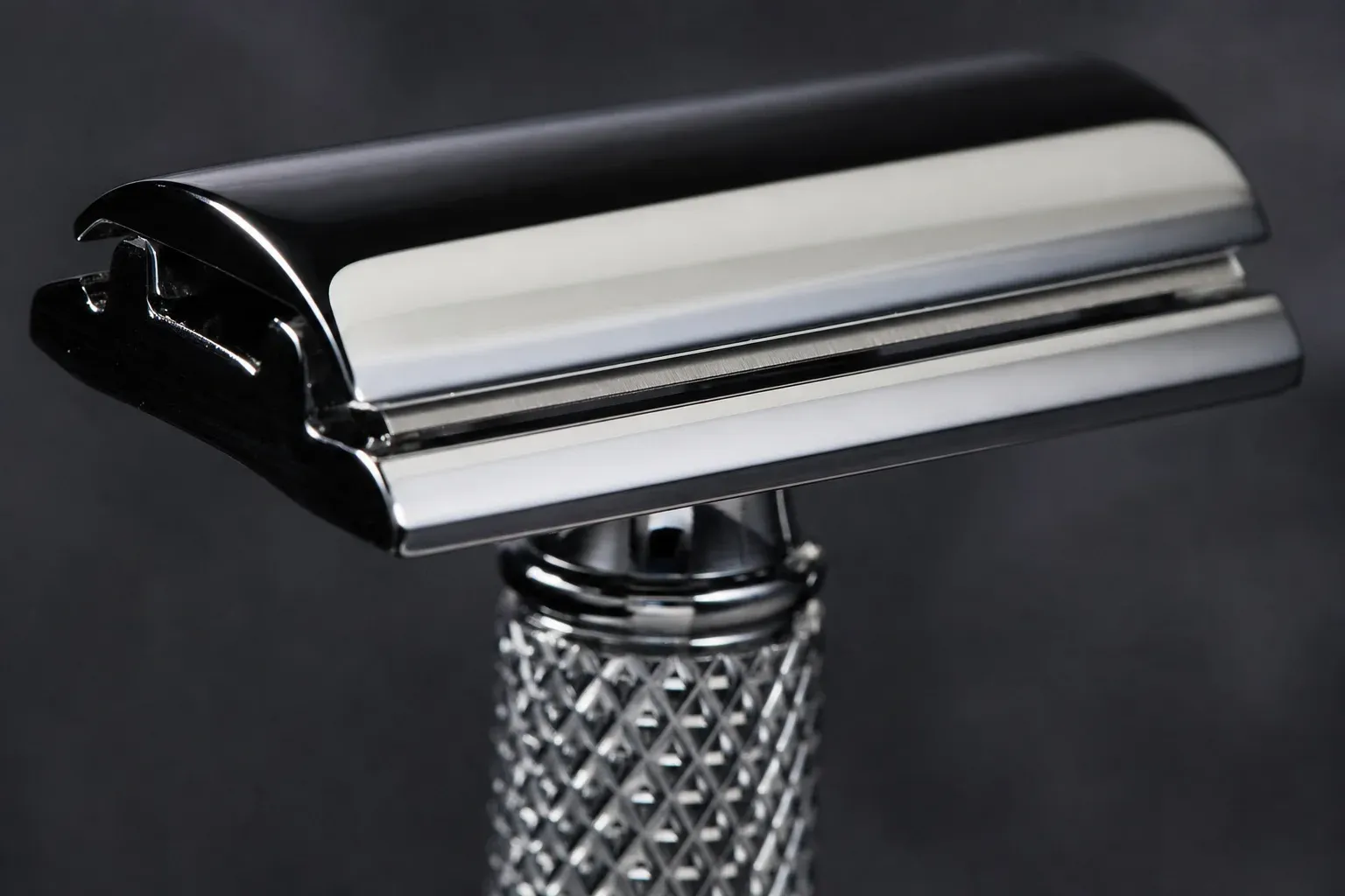 Macro photograph of a chrome closed-comb double-edge razor head against dark slate, the domed top cap over the visible blade edge and an unbroken smooth safety bar with no teeth, knurled handle below