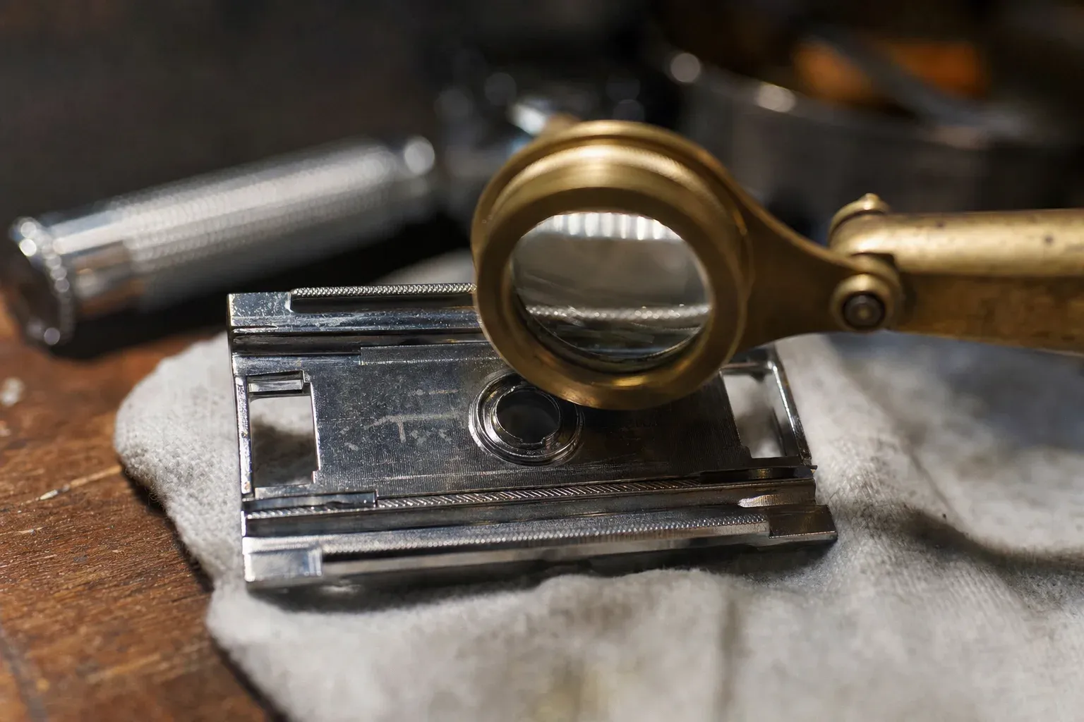 A small brass loupe resting on the underside baseplate of a disassembled vintage chrome safety razor as if inspecting it, with faint worn stamped markings on the metal, shallow depth of field under warm workbench light