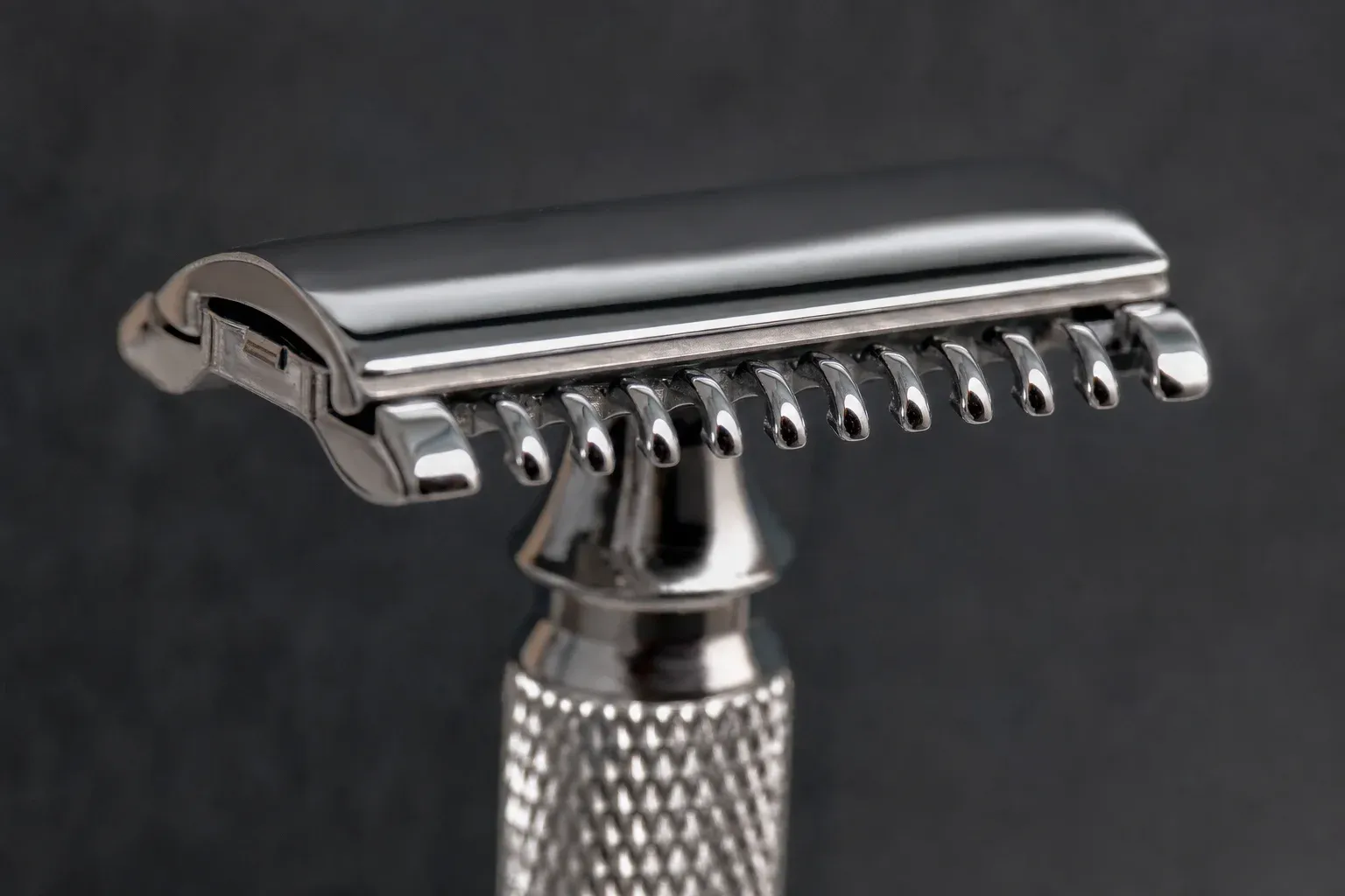 Macro photograph of an open-comb double-edge razor head against dark slate, a row of rounded chrome comb teeth with open gaps beneath the blade edge instead of a solid bar, knurled handle below