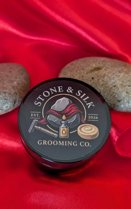 A Stone and Silk Grooming Co shave soap tub photographed from above on red satin between two smooth river stones, the lid showing the brand logo of a grey stone draped in red silk